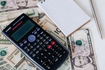 From above electronic calculator and notepad placed over United States dollar bills together with metallic pen for budget planning and calculation
