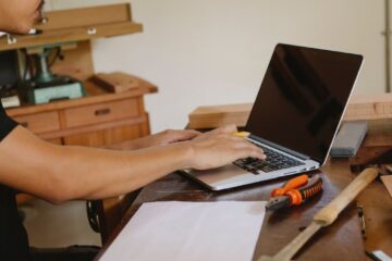 Crop unrecognizable man sitting at table with tools and browsing netbook while working in workshop