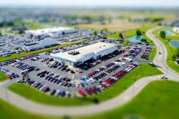 Aerial high-angle view of a bustling car dealership surrounded by parked cars in a green landscape.