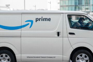 A white delivery van with Prime logo parked on an urban street with driver inside.