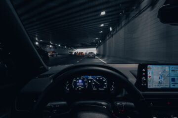 View from car dashboard driving through an illuminated city tunnel at night.
