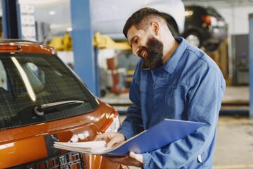 Smiling auto mechanic inspects a car in a service garage, wearing coveralls and holding documents.