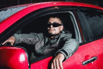 Man wearing sunglasses sitting in a red car during a rainy night.